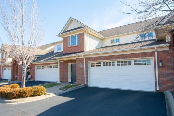 Brick attached Condominium with a tour car garage and small driveway. Winter time trees in front of the house with a beautiful blue sky